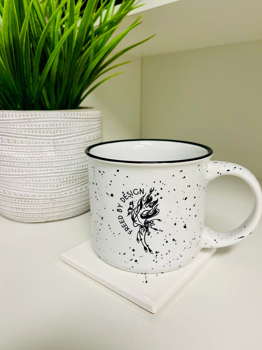 White mug with black speckles and design on a white surface next to a potted plant.